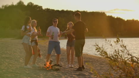 Company-of-five-young-people-are-dancing-in-shorts-and-t-shirts-around-bonfire-on-the-open-air-party-with-beer.-They-are-clink-and-drink-beer-and-enjoying-the-summer-evening-at-sunset-near-the-forest.